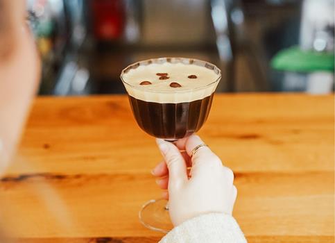 Eine Hand hält ein Glas mit cremigem Espresso Martini, garniert mit Kaffeebohnen, an einer Bar.