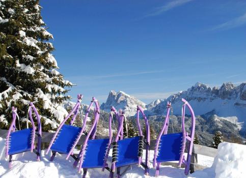 Mehrere bunte Rodeln im Schnee mit Blick auf verschneite Dolomiten und blauen Himmel.