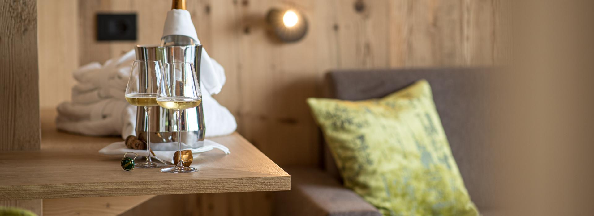 Zwei Gläser Weißwein mit Sektkühler auf einem Holztisch vor einer gemütlichen Couch mit einem grünem Kissen.