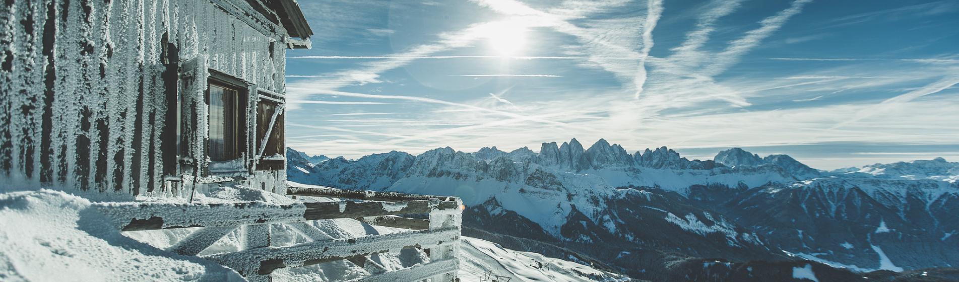 Snow-covered, icy mountain hut with view of the Dolomites and bright winter sun.