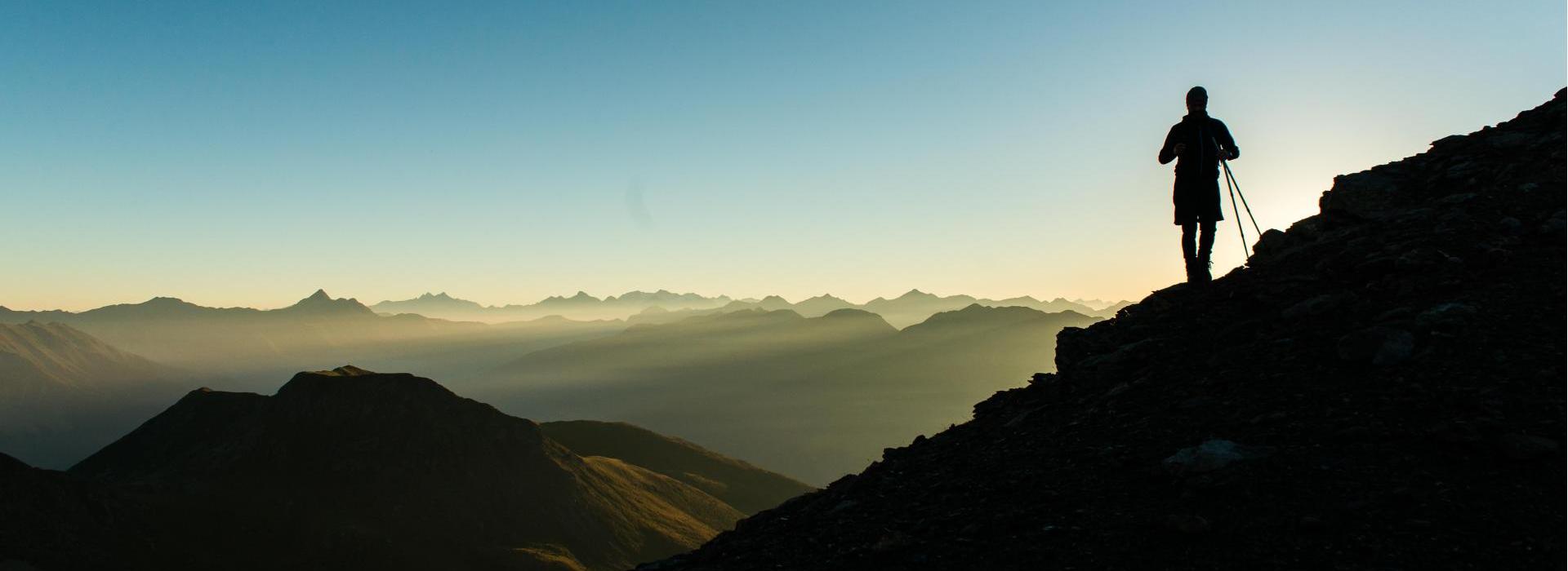 Sagoma di un escursionista all’alba su un pendio montano con vista sulla catena montuosa.