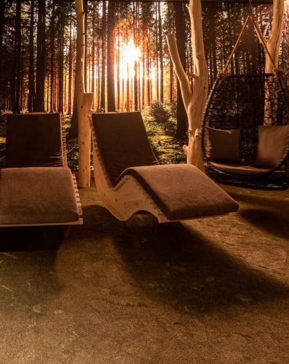 Sala relax con lettini in legno e sedia sospesa davanti a un'immagine di foresta al tramonto