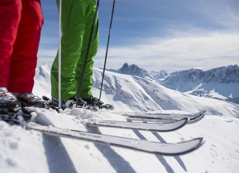 Zwei Skifahrer mit bunten Hosen stehen bereit auf verschneitem Gipfel mit Bergpanorama.