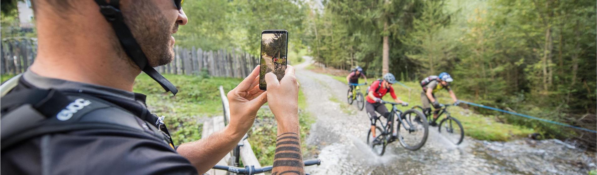 A mountain biker takes a photo of cyclists riding through a stream in the forest.