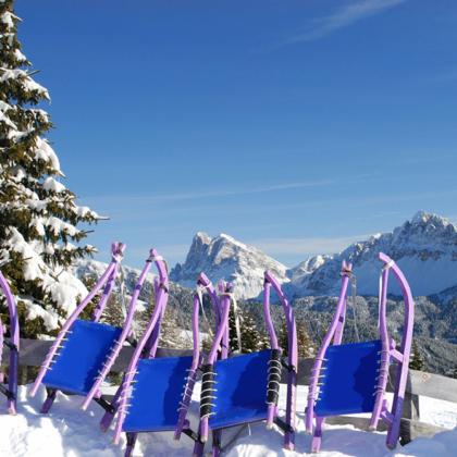 Mehrere bunte Rodeln im Schnee mit Blick auf verschneite Dolomiten und blauen Himmel.