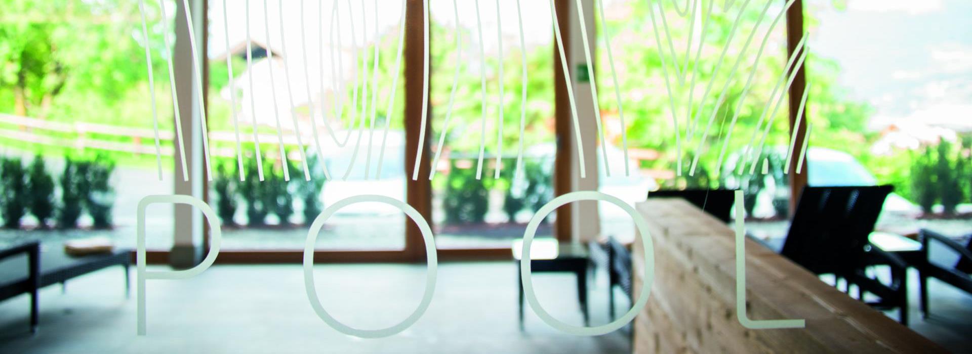 View through a glass wall with POOL lettering into a bright room with windows and loungers.