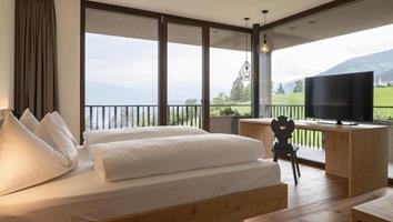 Camera luminosa con letto grande, TV e finestre panoramiche con vista sul paesaggio verde.