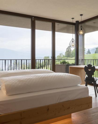 Bright hotel room with large bed, TV, and panoramic windows offering a view of the green landscape.