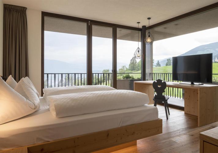 Helles Hotelzimmer mit großem Bett, TV und Panoramafenstern mit Ausblick auf grüne Landschaft.
