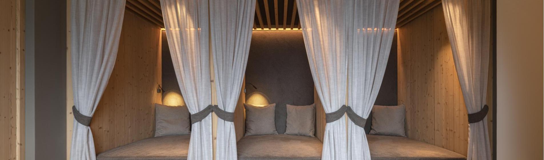 Zona relax accogliente con tre lettini, tende, soffitto in legno e luce d’atmosfera.