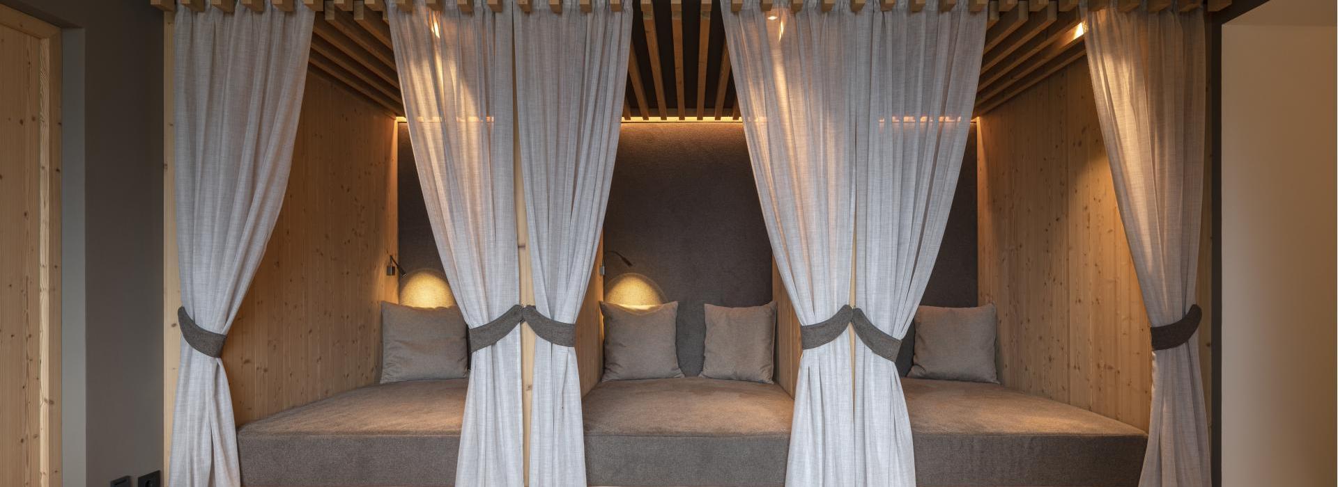 Zona relax accogliente con tre lettini, tende, soffitto in legno e luce d’atmosfera.