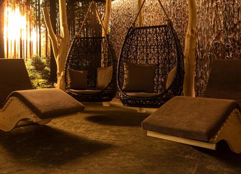 Sala relax accogliente con lettini in legno e sedute sospese, illuminata da luce calda e naturale.