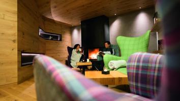 Gemütliche Lounge mit Kamin, zwei Gäste lesen entspannt in bunten Sesseln bei Kerzenschein.