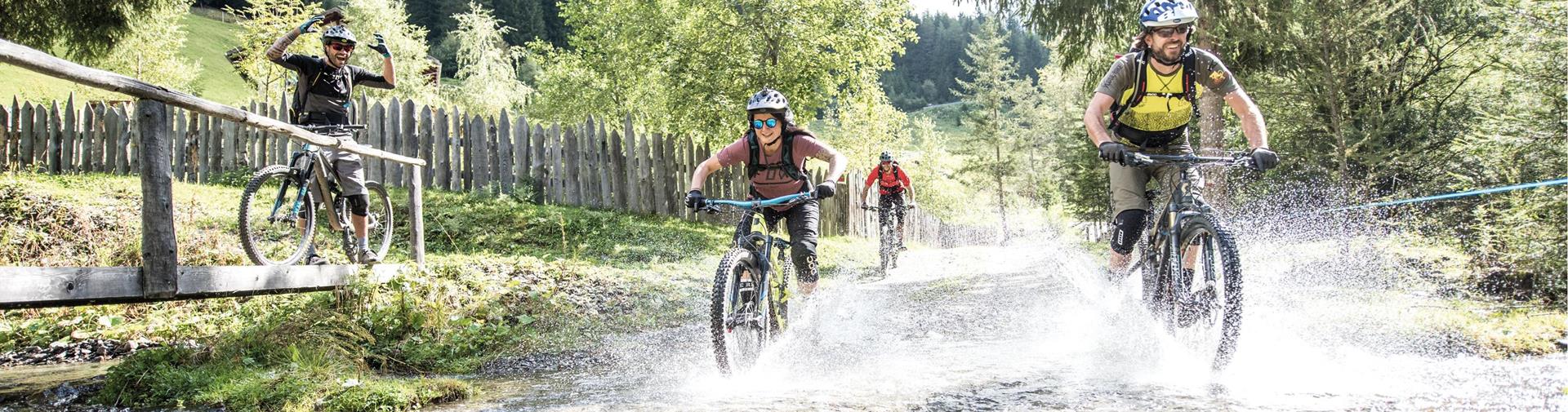 Mountainbiker durchqueren lachend einen Bach im Wald, während ein Fahrer jubelnd zuschaut.