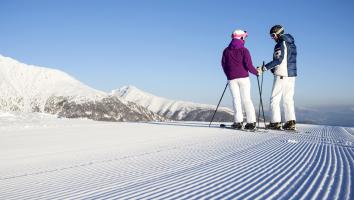 Two skiers on a freshly groomed slope are enjoying the view of snow-covered mountain peaks.