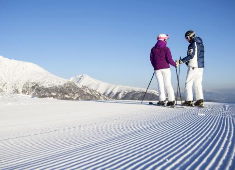 Two skiers on a freshly groomed slope are enjoying the view of snow-covered mountain peaks.