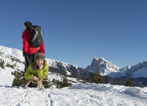 Child smiling on a sled, pulled by an adult, on snowy ground with scenic mountain view.