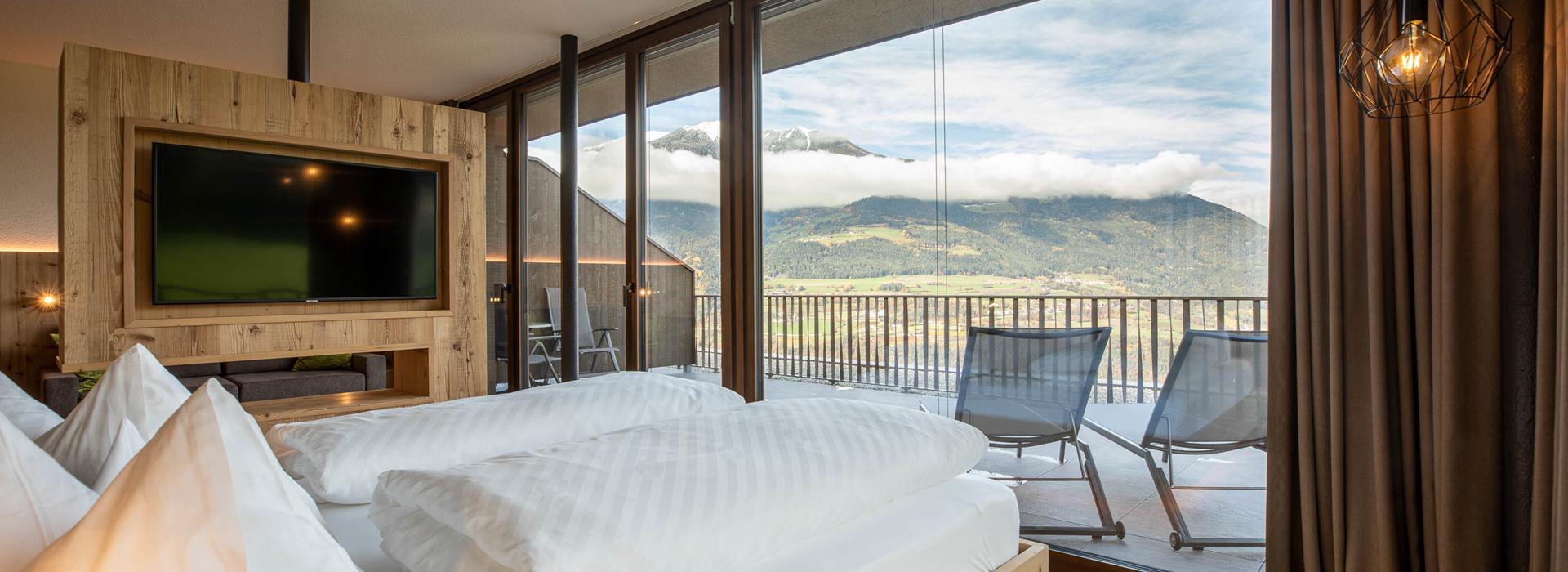 Modernes Hotelzimmer mit Holzelementen, Panoramafenstern, großem Bett und Bergblick vom Balkon.