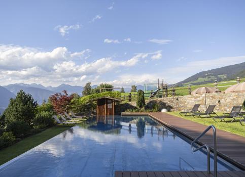 Piscina esterna con lettini prendisole, panorama montano e giardino verde in una giornata di sole.