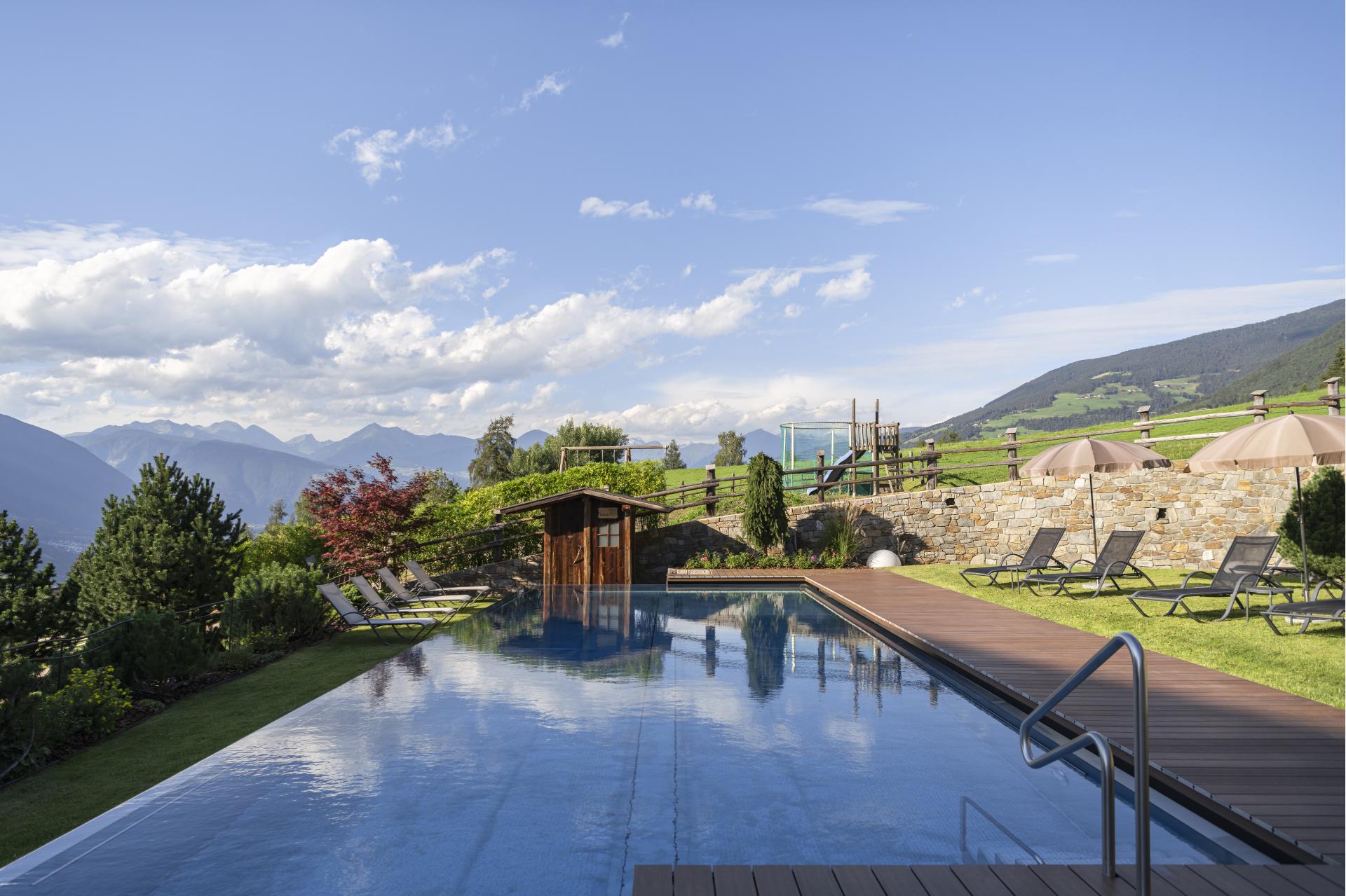 Outdoor pool with sun loungers, mountain panorama, and green garden on a sunny day.