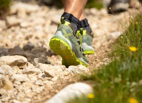 Close-up of hiking shoes on a rocky mountain trail with grass and wildflowers along the edge.