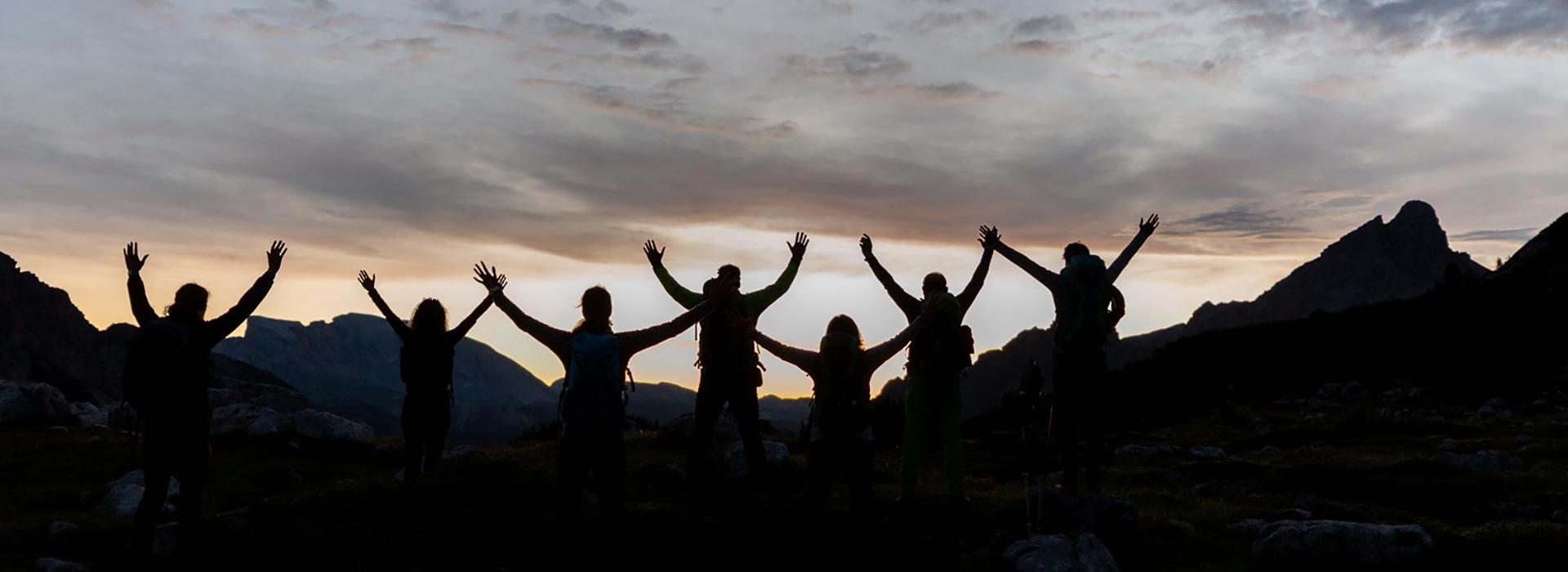 Silhouetten einer Gruppe von Menschen mit erhobenen Armen vor einem dramatischen Sonnenuntergang in den Bergen.