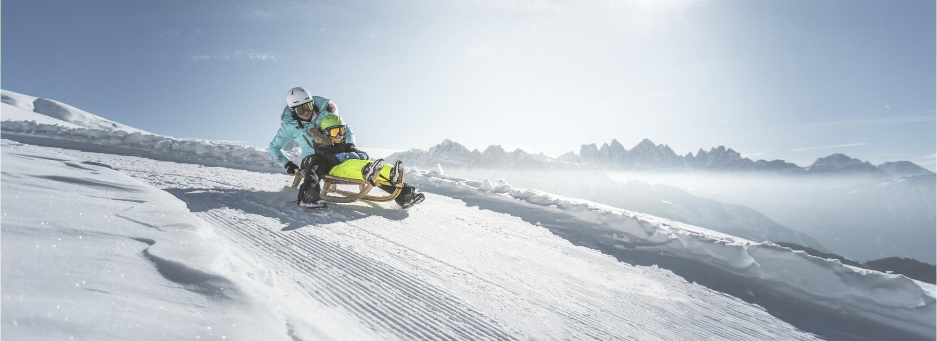 Two people sled down a snowy slope in the sun with a scenic view of the Dolomites in the background.
