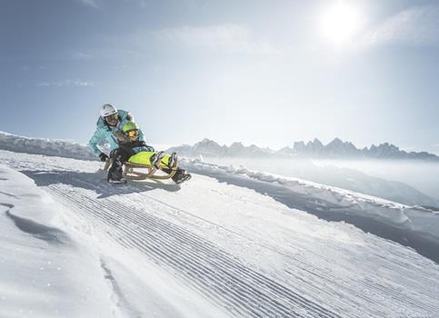 Two people sled down a snowy slope in the sun with a scenic view of the Dolomites in the background.