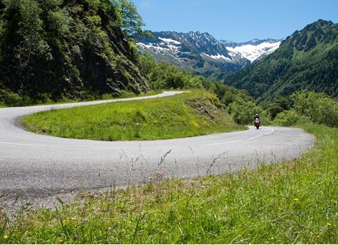Motorcyclist on a winding alpine road surrounded by green hills and snow-capped mountains.