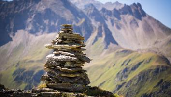 Un mucchio di pietre impilate artisticamente si erge su un altopiano alpino con montagne sullo sfondo.
