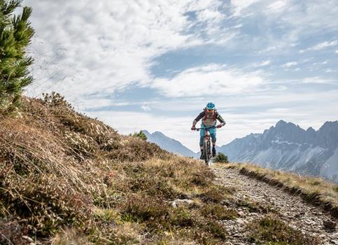 Ein Mountainbiker fährt auf einem schmalen Pfad durch die alpine Berglandschaft mit felsigen Gipfeln.