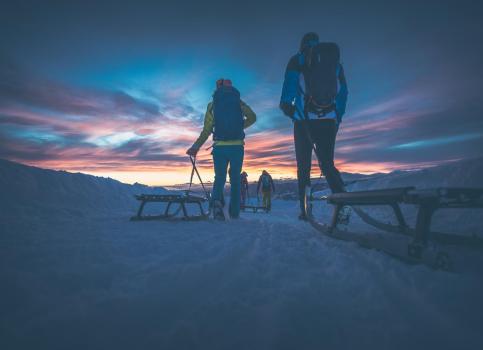 People pulling sleds on snowy trail at sunset beneath a vivid, colorful sky.