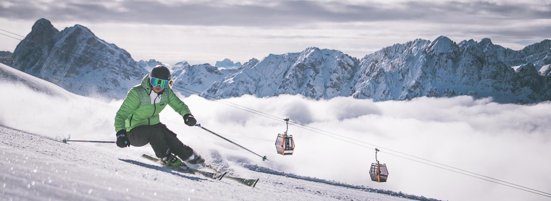 Skier carving on fresh slope, cable cars and snowy Dolomites in the background.