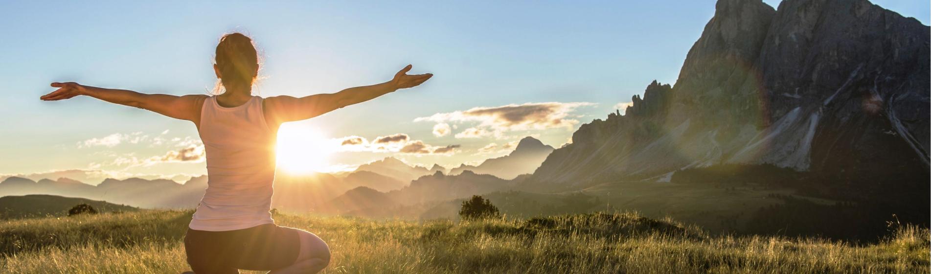 Woman kneeling with open arms at sunrise in front of a breathtaking mountain landscape.